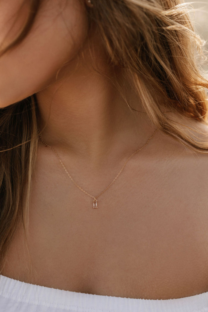 A close-up of a woman's neck wearing the LLUME Jewelry Whisper Necklace—a gold-plated sterling silver piece with a small rectangular pendant—against her light skin, white off-shoulder top, and partially visible loose hair.