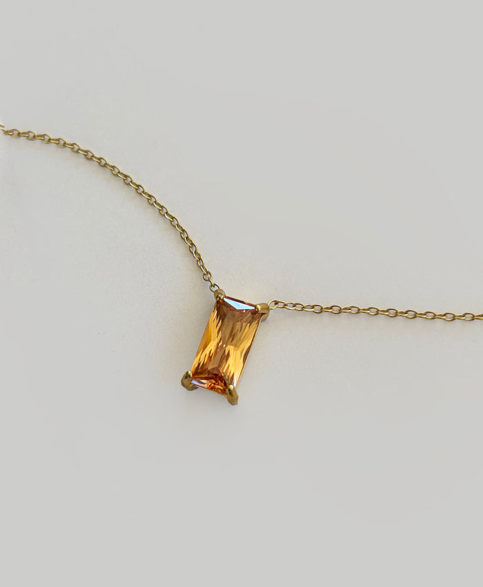 The Wild Honey Necklace by LLUME Jewelry features a rectangular, amber-colored gemstone pendant—reminiscent of honey—set in a simple gold prong setting, displayed against a light gray background.