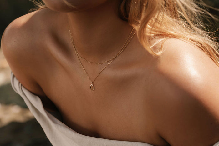 A close-up shows a woman in an off-the-shoulder light top, her wavy blonde hair to one side, elegantly wearing the Wishbone Daily Set by LLUME Jewelry.