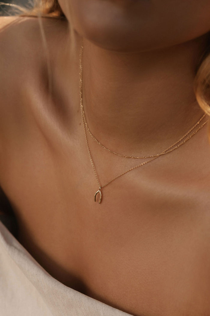 A close-up of a light-skinned woman in off-shoulder cream fabric wearing the LLUME Jewelry Wishbone Daily Set—a delicate gold necklace set featuring a small Wishbone Necklace pendant—captured in soft, warm lighting.