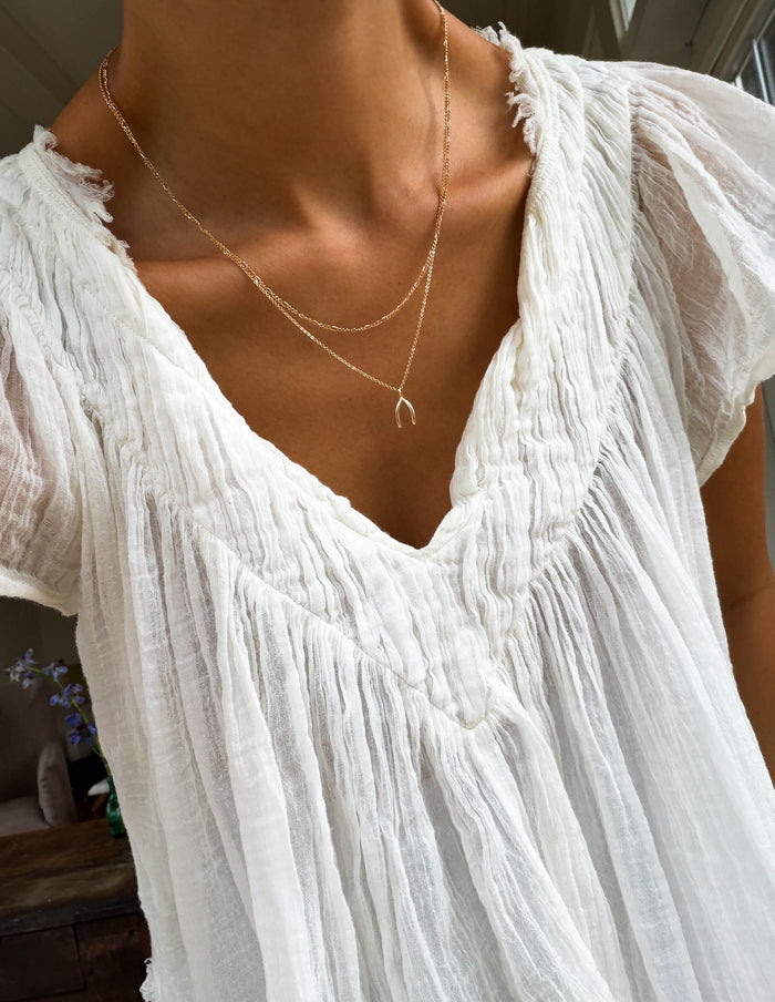 A person wears the Wishbone Daily Set by LLUME Jewelry with a sheer, white, textured V-neck blouse. The photo is cropped to show only the neck, chest, and upper torso.