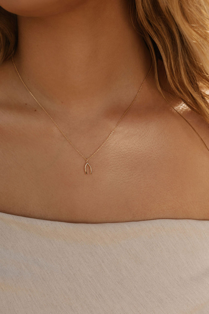 A woman wears the Wishbone Necklace by LLUME Jewelry, featuring a delicate wishbone pendant; her wavy hair and light off-shoulder top showcase this versatile accessory.