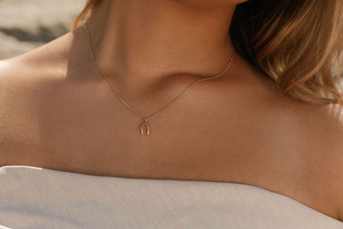 A woman exudes timeless elegance in an off-the-shoulder light top, highlighted by the LLUME Jewelry Wishbone Necklace, its delicate gold charm softly illuminated by natural light.