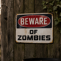 Close up of a Beware Of Zombies metal warning sign on a weathered wooden fence with zombie figures in the background
